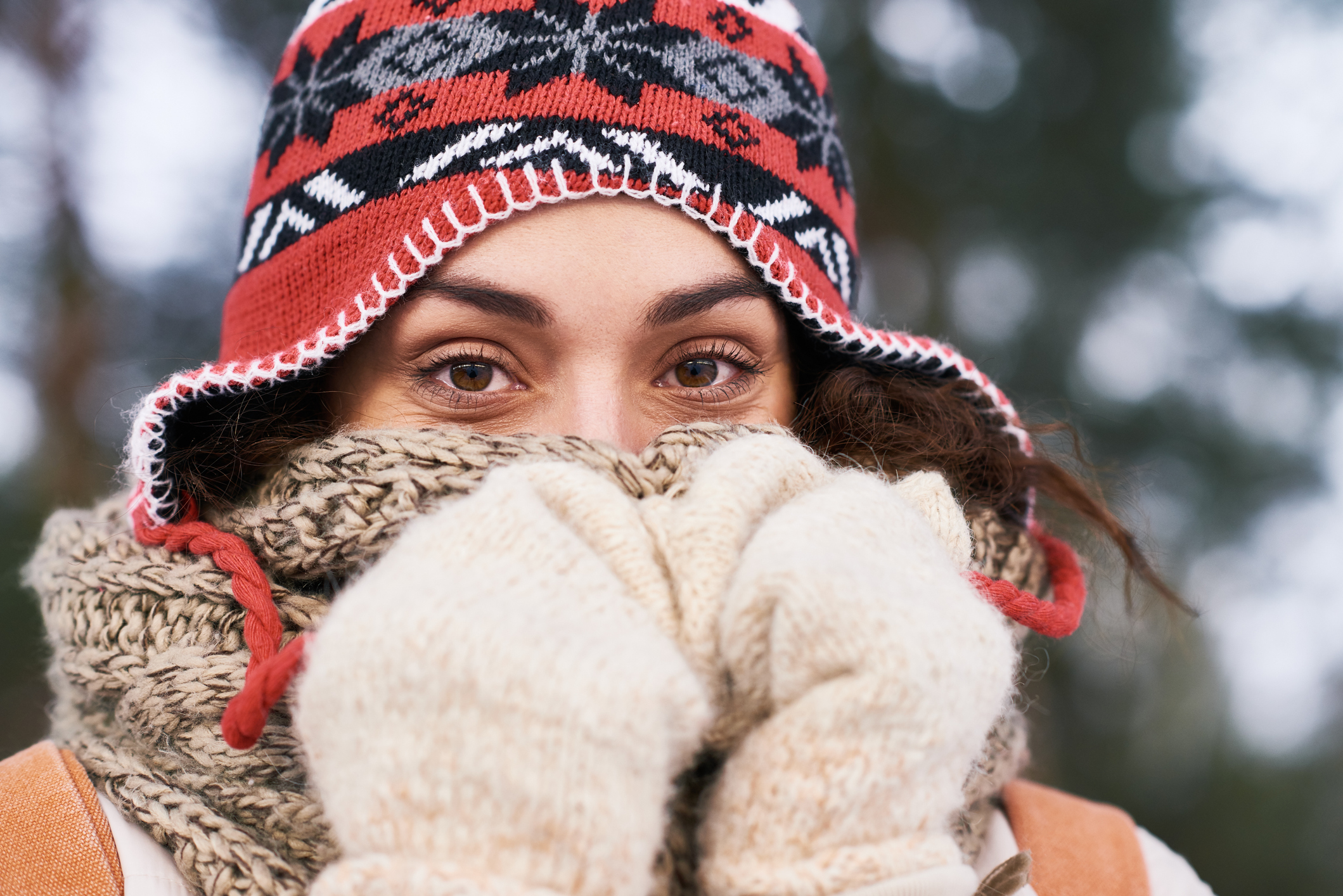 Close-up of beautiful young woman covering nose with wool scarf and mittens looking at camera outdoors