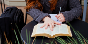 A young, red-haired woman writing notes as she reads the Bible. She is sitting at a cafe with her suitcase next to her because she is traveling.