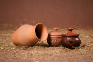 Clay jug, and pots with lids on the hay on a brown background in the studio, horizontal photo