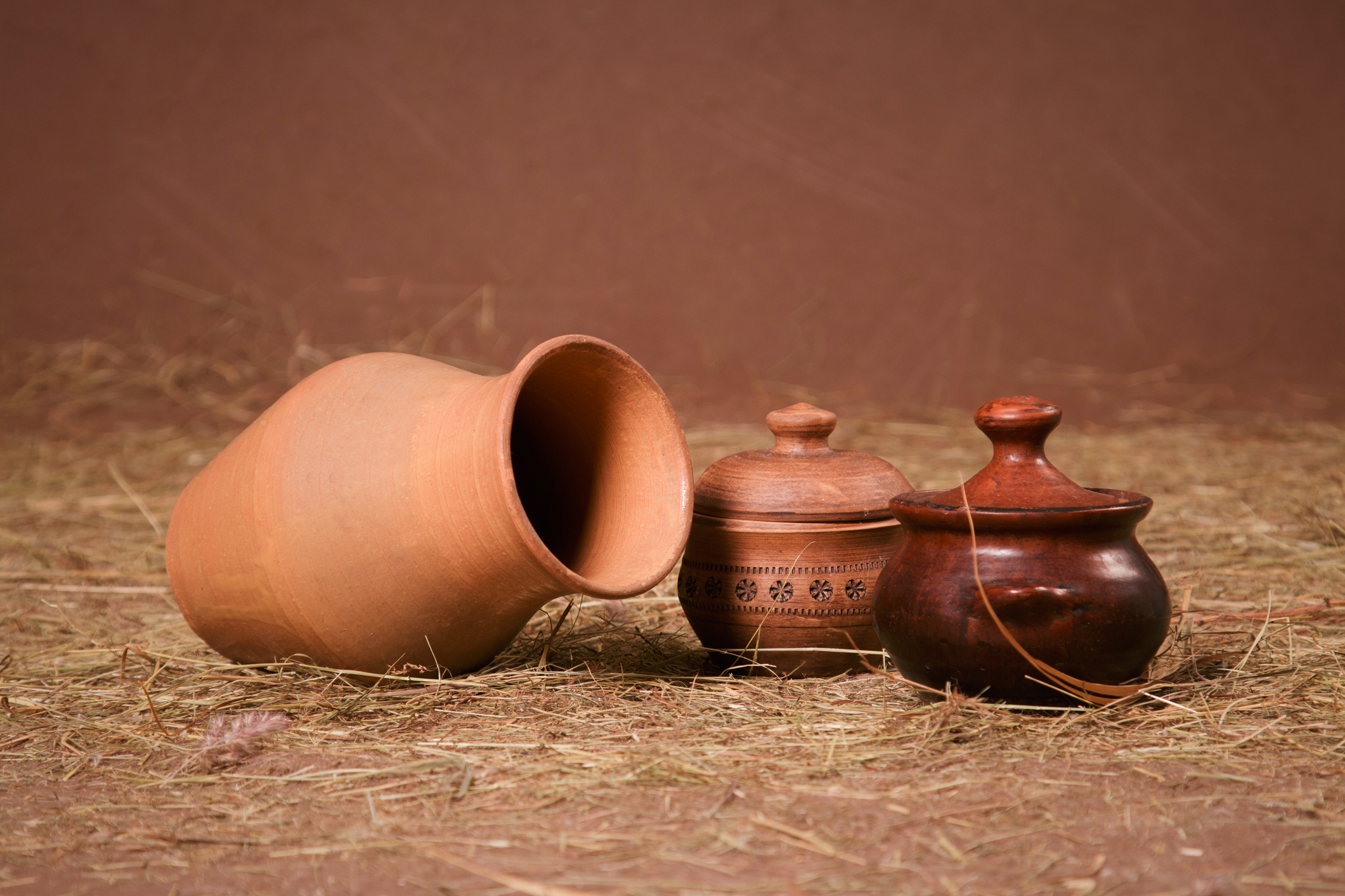 Clay jug, and pots with lids on the hay on a brown background in the studio, horizontal photo